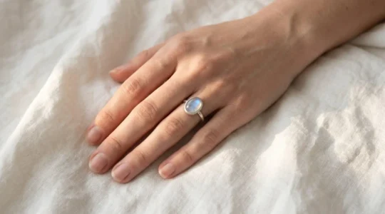 Gros plan d'une main féminine portant une bague en pierre de lune ovale sur argent 925, posée sur du lin blanc, lumière naturelle révélant les reflets bleutés de la pierre