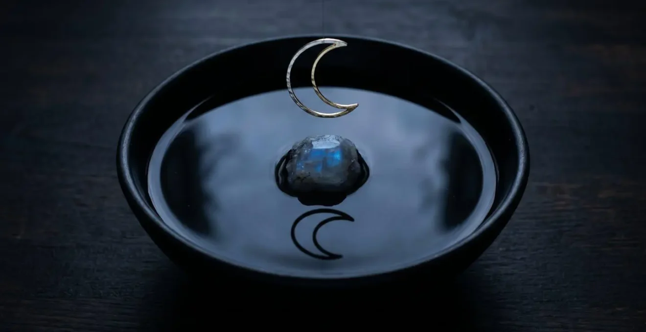 Pierre de lune cabochon posée sur un miroir d'eau sombre avec croissant de lune doré suspendu au-dessus, ambiance nocturne poétique